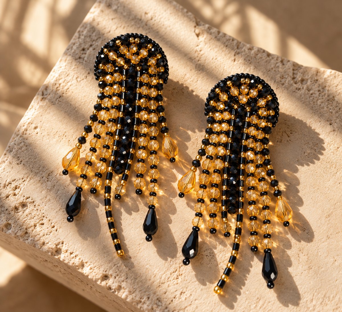 Par de aretes Medusa negros y dorados con flecos y colgantes en forma de gota sobre piedra clara.