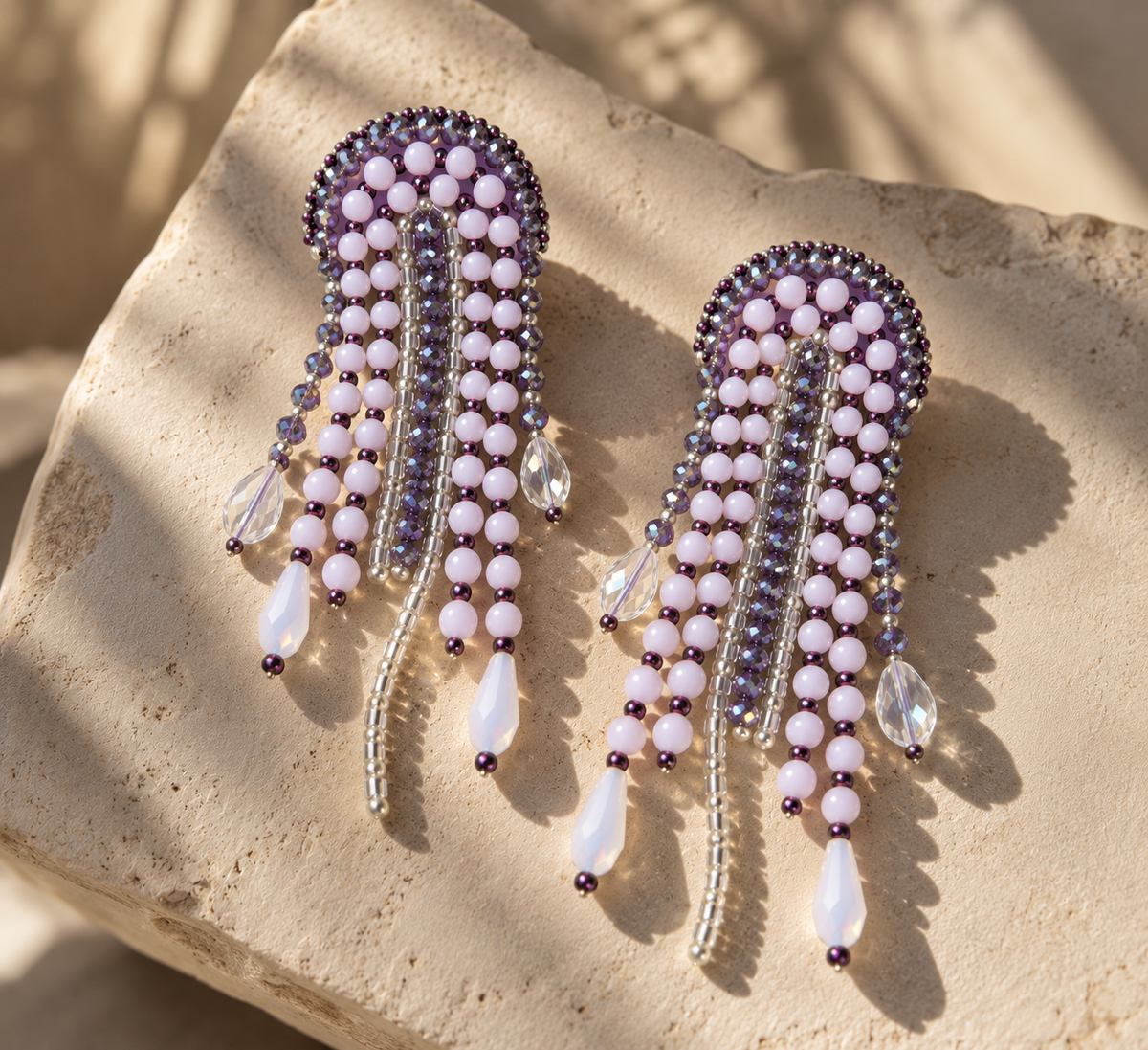 Par de aretes Medusa lilas con cuentas y colgantes en forma de gota sobre piedra al sol.