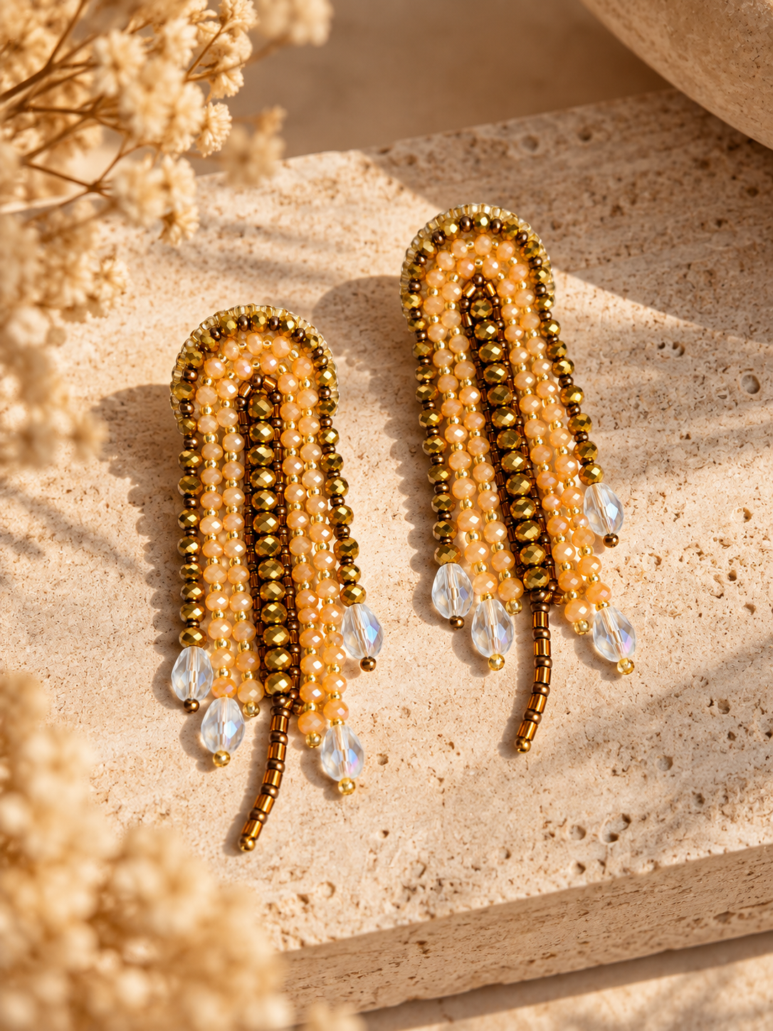 Par de aretes Medusa dorados con cuentas claras y colgantes transparentes sobre piedra iluminada por el sol.