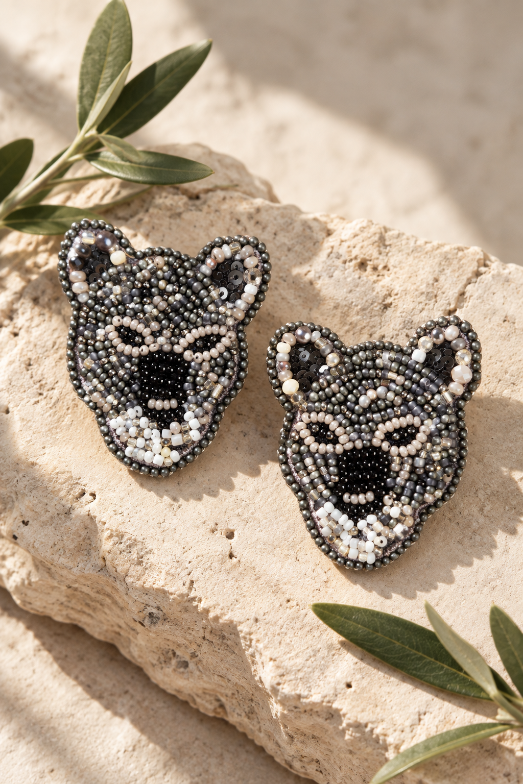Par de aretes Leopardo plateados de SALÁ, con cuentas grises, negras y blancas sobre piedra beige junto a hojas verdes.