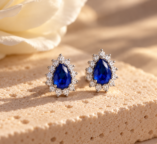Par de aretes Coral Azul con piedras azules en forma de gota y halo de piedras transparentes, sobre piedra beige.