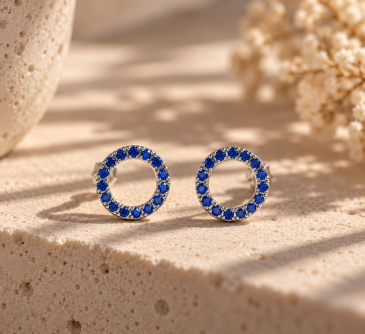 Aretes Mona Azul de diseño circular con centro abierto y pequeñas piedras azules, sobre piedra beige.