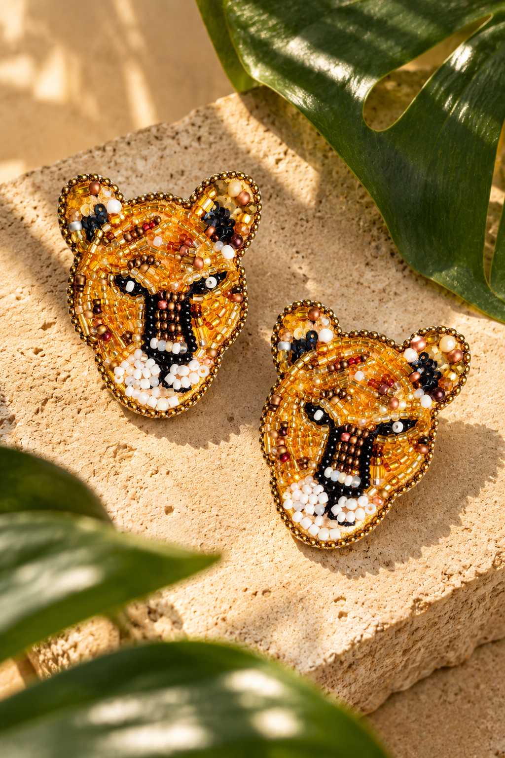 Aretes Leopardo dorados de SALÁ en forma de rostro felino, con detalles negros y blancos sobre piedra junto a hojas verdes.