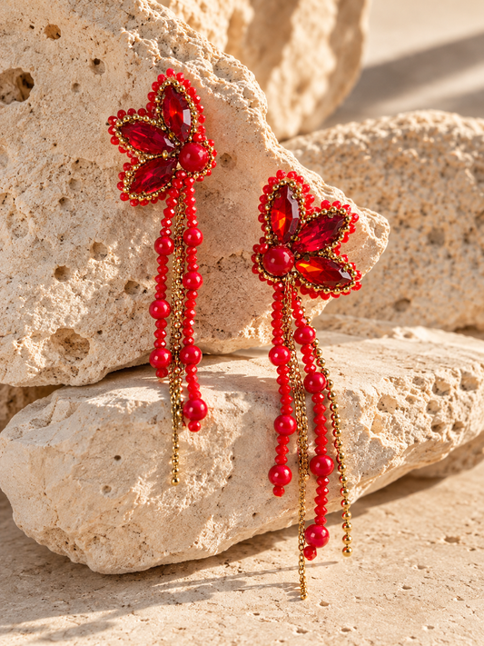 Aretes Cayena rojos con cristales en forma de pétalos, cuentas colgantes y cadenas doradas sobre piedra beige.