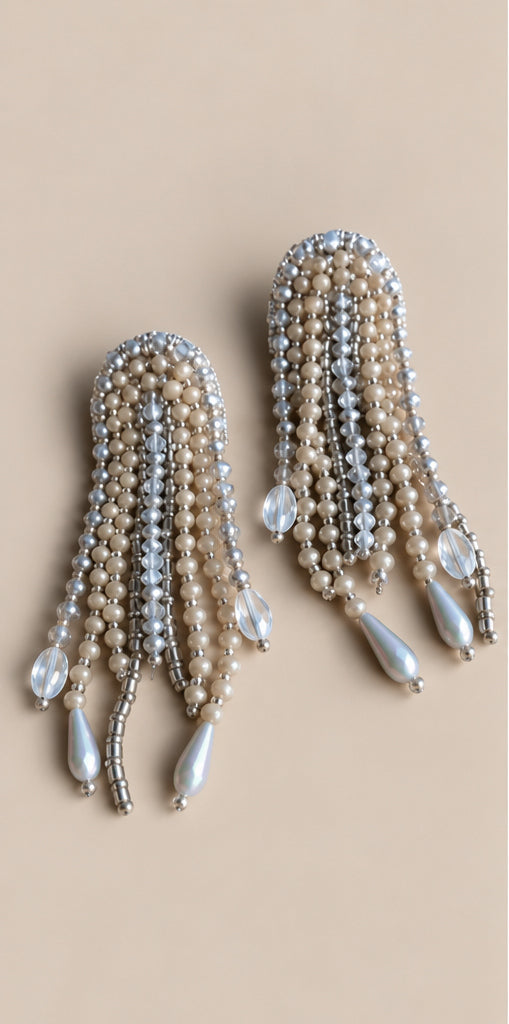 Par de aretes Medusa plateados con cuentas claras y colgantes en forma de gota sobre fondo beige.